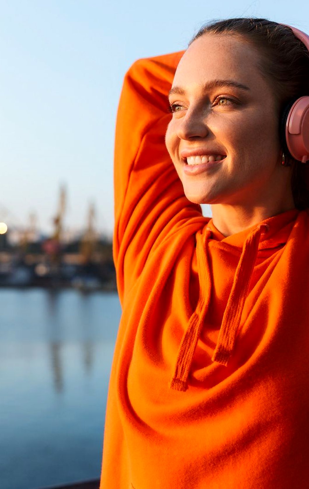 Smiling woman in an orange hoodie and headphones by the water