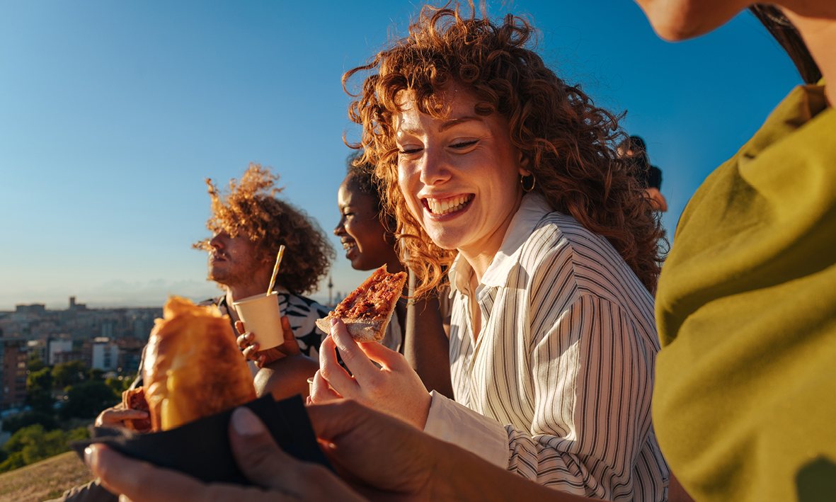 Friends laughing and sharing food outdoors on a sunny day, with the city behind them